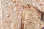 A close-up view of a delicate, beige-colored fabric with intricate gold and white sequin designs, including floral and leaf patterns. The fabric appears to be a sheer or sheer-like material, with a sheer overlay.