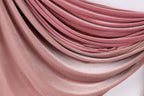 a close-up view of a fabric with a soft, flowing texture in shades of pink and red. The fabric appears to be made of a soft, plush material, possibly velvet or a similar type of material. The fabric is draped or folded in a way that creates a sense of movement and depth, with the colors gradually transitioning from a lighter shade at the top to a darker shade at the bottom.