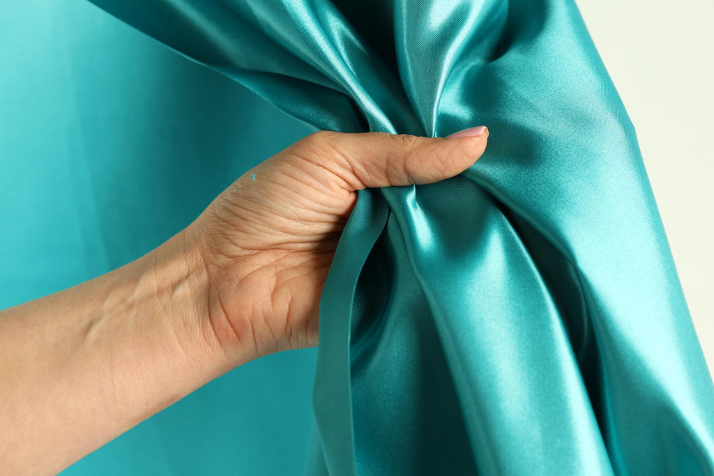 A hand is holding a turquoise satin curtain, partially covering it.