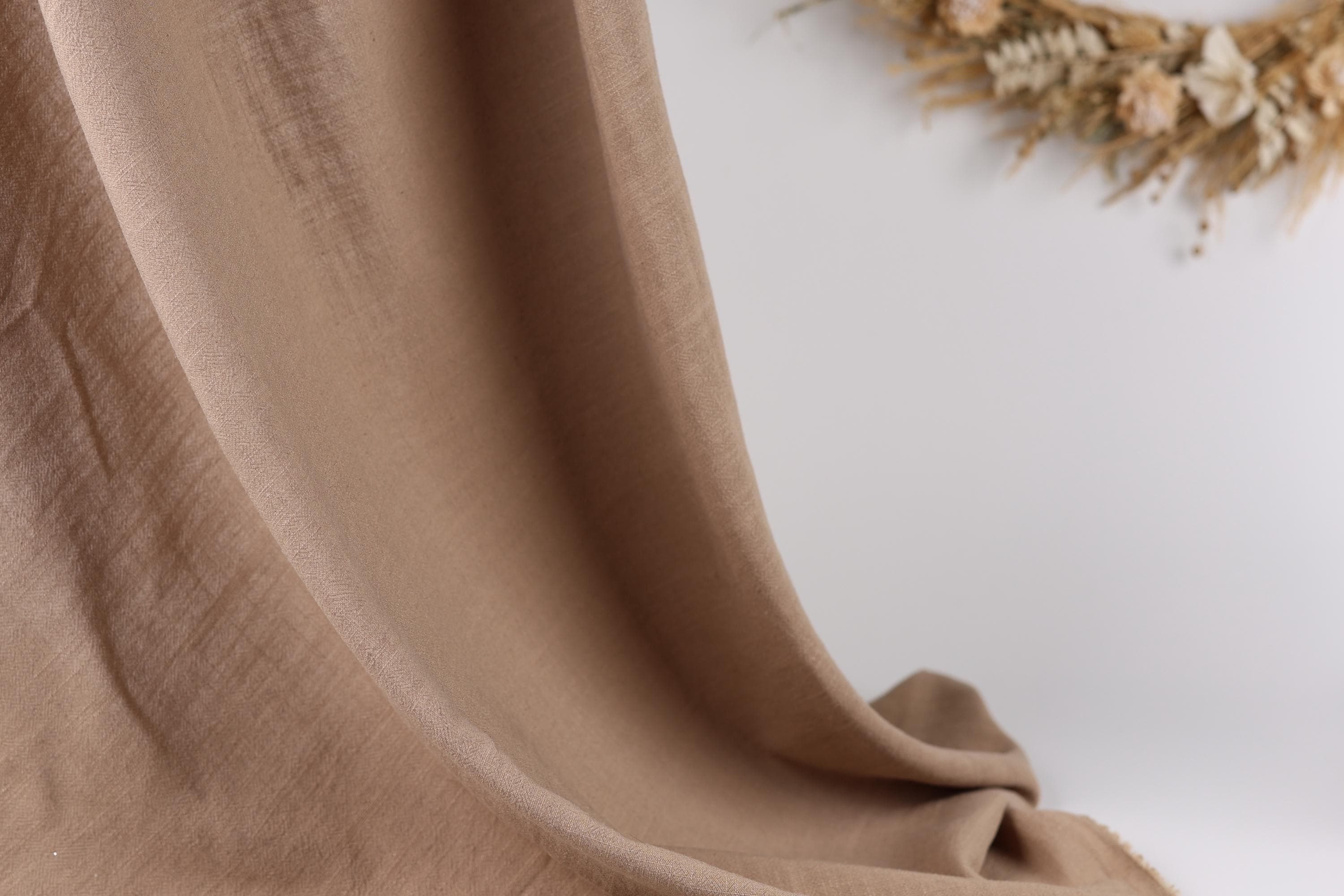 a close-up view of a beige fabric, possibly a piece of clothing or a textile, with a blurred background that includes dried flowers.
