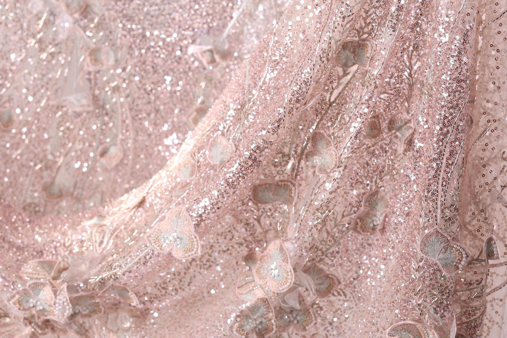 The image is a close-up of a delicate, pink, and beige fabric with a shimmering, sequined texture. The fabric appears to be made of lace or a similar delicate material, with a floral pattern visible on the surface. The image does not contain any text or other discernible objects or actions.