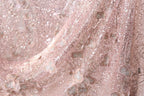 The image is a close-up of a delicate, pink, and beige fabric with a shimmering, sequined texture. The fabric appears to be made of lace or a similar delicate material, with a floral pattern visible on the surface. The image does not contain any text or other discernible objects or actions.