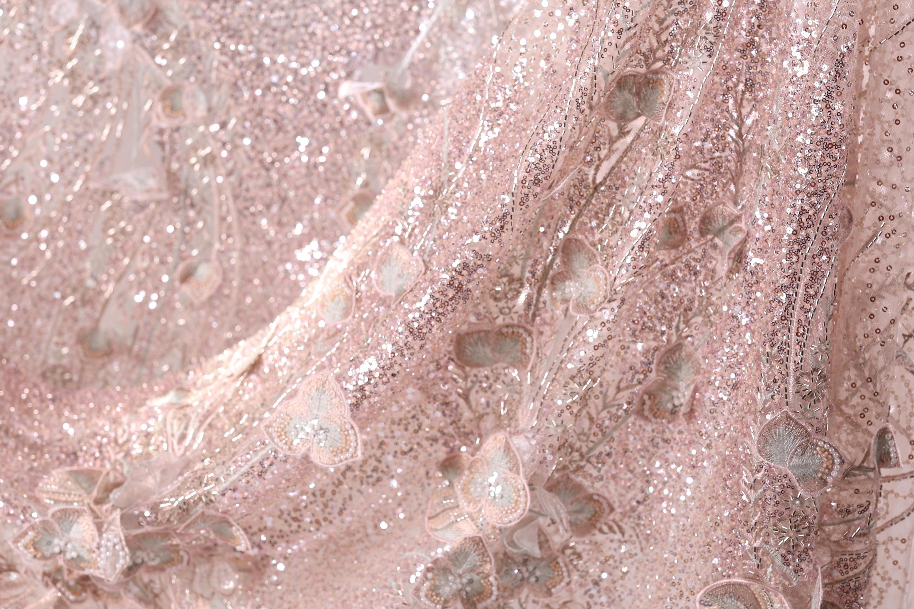 The image is a close-up of a delicate, pink, and beige fabric with a shimmering, sequined texture. The fabric appears to be made of lace or a similar delicate material, with a floral pattern visible on the surface. The image does not contain any text or other discernible objects or actions.