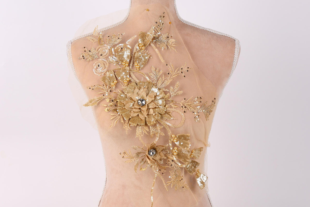 a mannequin with a back adorned with a delicate, gold-colored floral design. The design includes a large flower at the center, surrounded by smaller flowers and leaves, all intricately crafted with gold sequins and beads. The mannequin is positioned against a plain white background, allowing the gold floral design to stand out prominently.