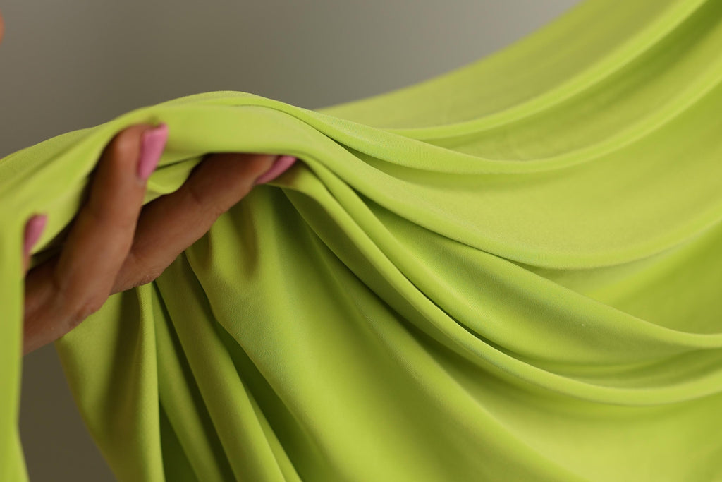 A hand is holding a bright green fabric, which appears to be a soft, flowing material.