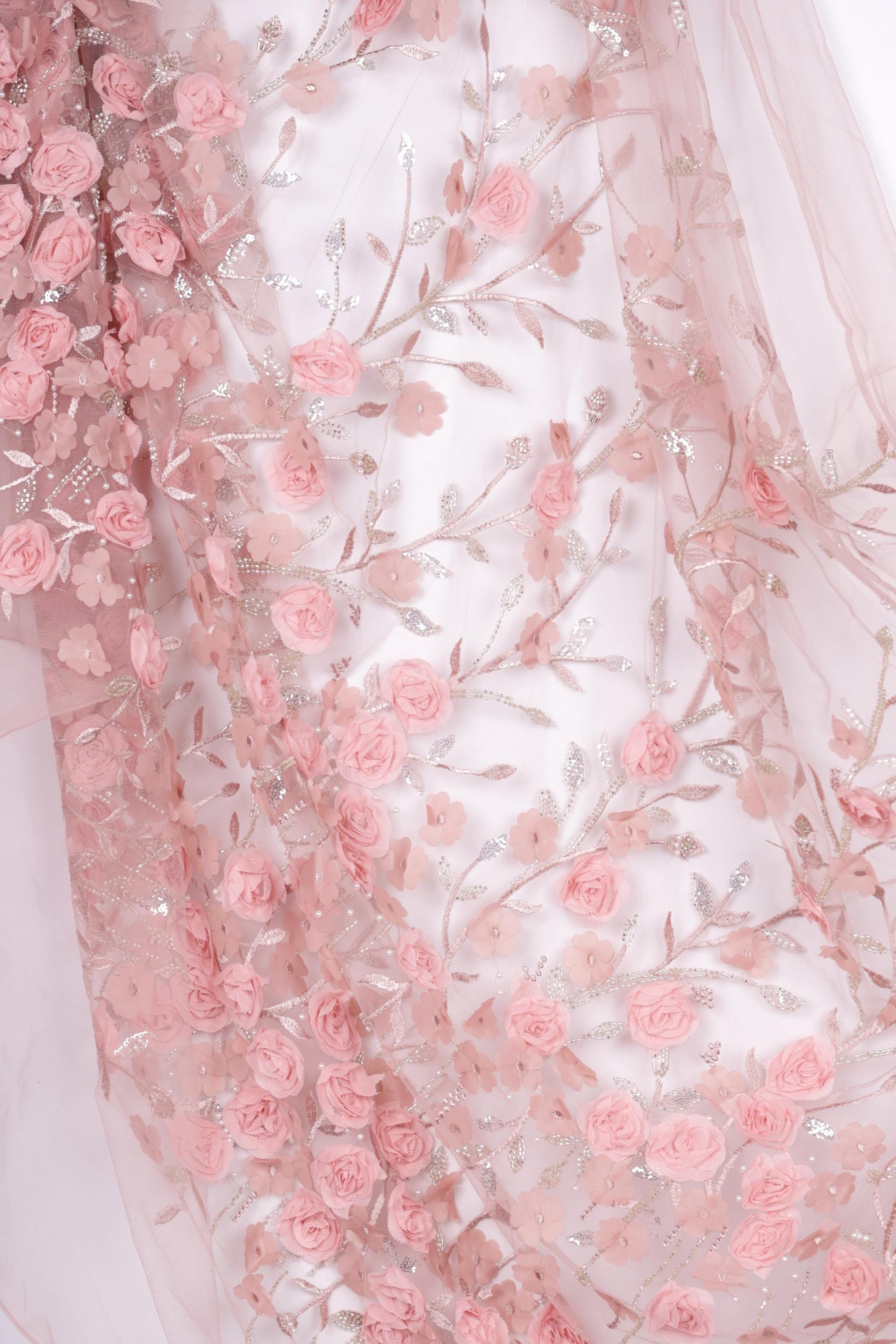 a close-up view of a delicate, pink floral fabric with numerous small pink roses and leaves. The fabric appears to be made of sheer material, giving it a soft and delicate appearance. The roses and leaves are arranged in a way that creates a visually appealing and intricate pattern.