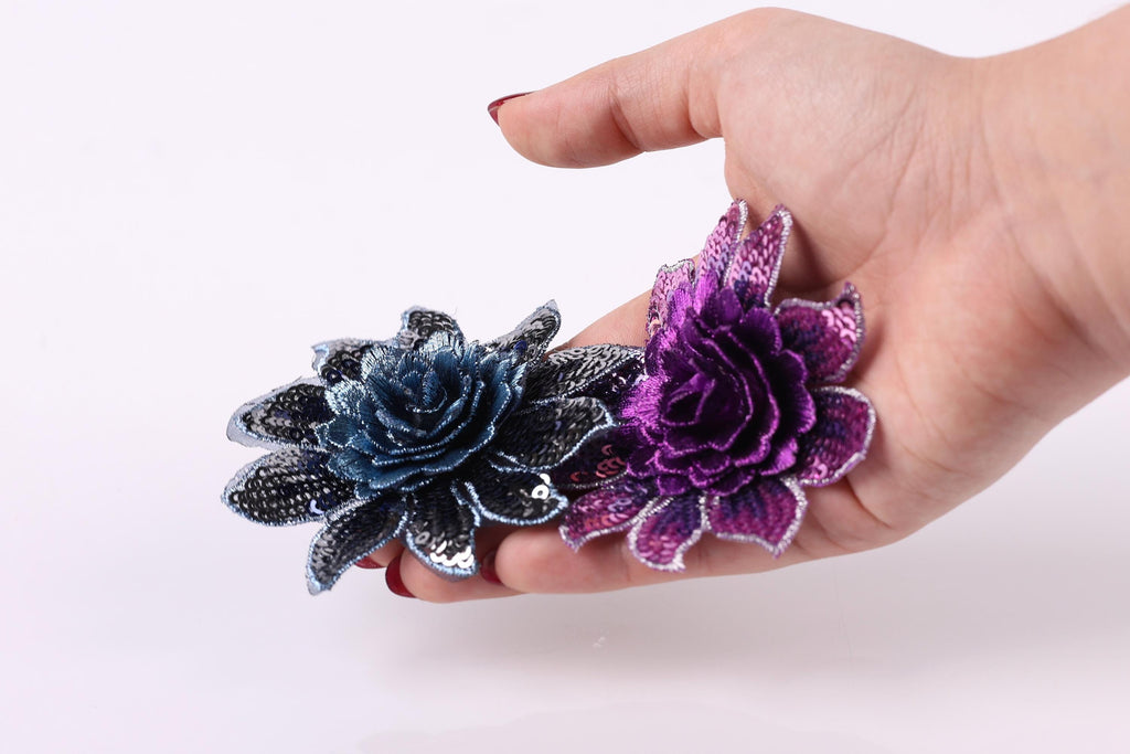A hand holding two colorful flower-shaped objects, one blue and one purple, against a plain white background.