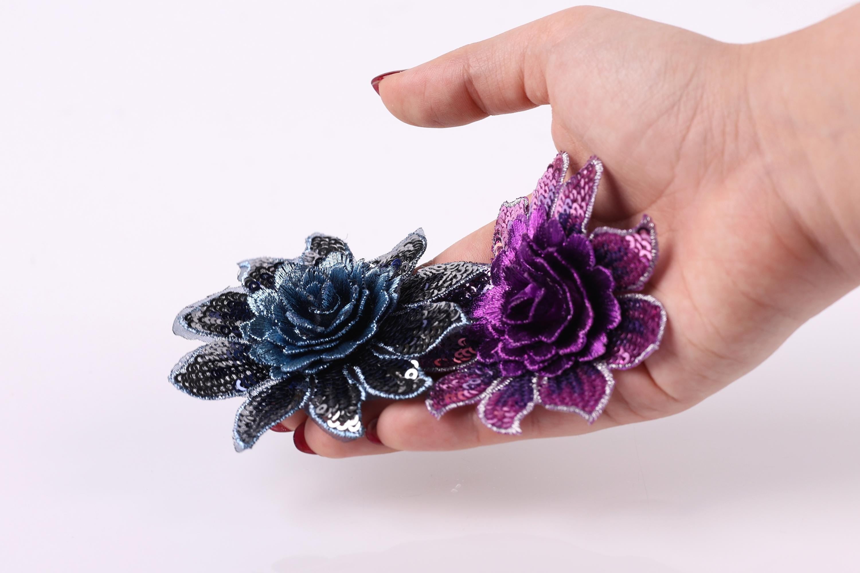 A hand holding two colorful flower-shaped objects, one blue and one purple, against a plain white background.