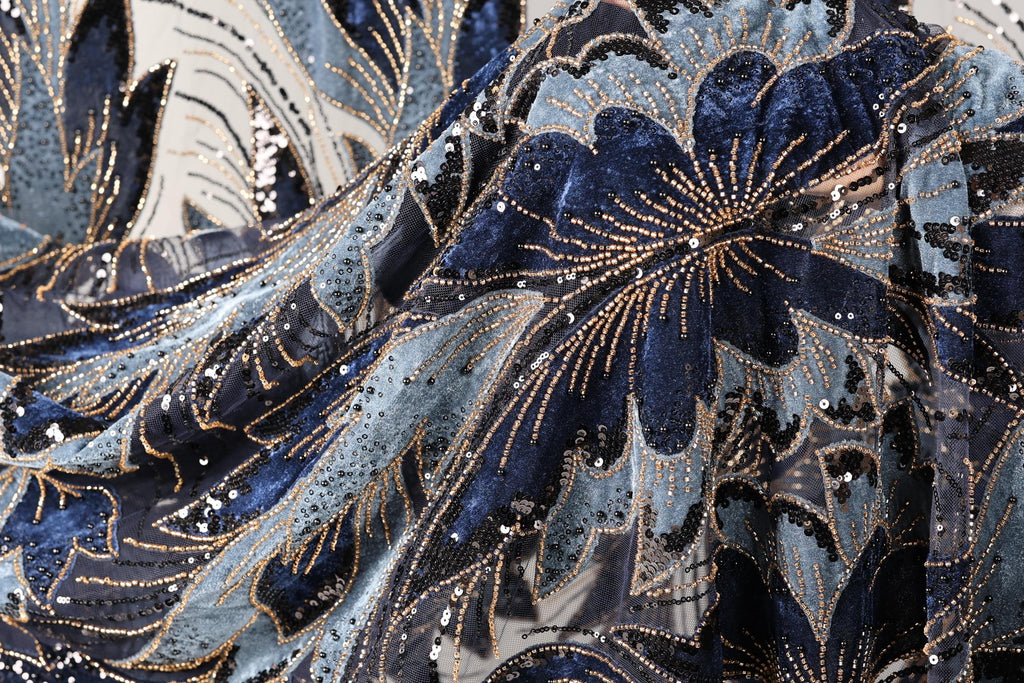 The image is a close-up of a fabric with a floral pattern, featuring blue and black colors and numerous sequins or beads. The fabric appears to be made of a shiny material, possibly velvet or a similar type of material. The intricate design and the presence of sequins give the fabric a luxurious and elegant appearance.