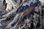 The image is a close-up of a fabric with a floral pattern, featuring blue and black colors and numerous sequins or beads. The fabric appears to be made of a shiny material, possibly velvet or a similar type of material. The intricate design and the presence of sequins give the fabric a luxurious and elegant appearance.