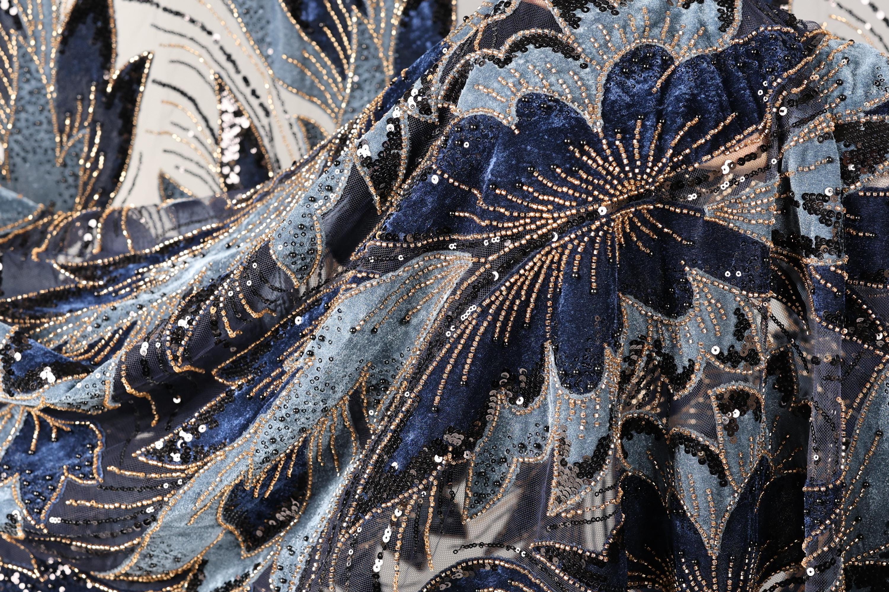 The image is a close-up of a fabric with a floral pattern, featuring blue and black colors and numerous sequins or beads. The fabric appears to be made of a shiny material, possibly velvet or a similar type of material. The intricate design and the presence of sequins give the fabric a luxurious and elegant appearance.