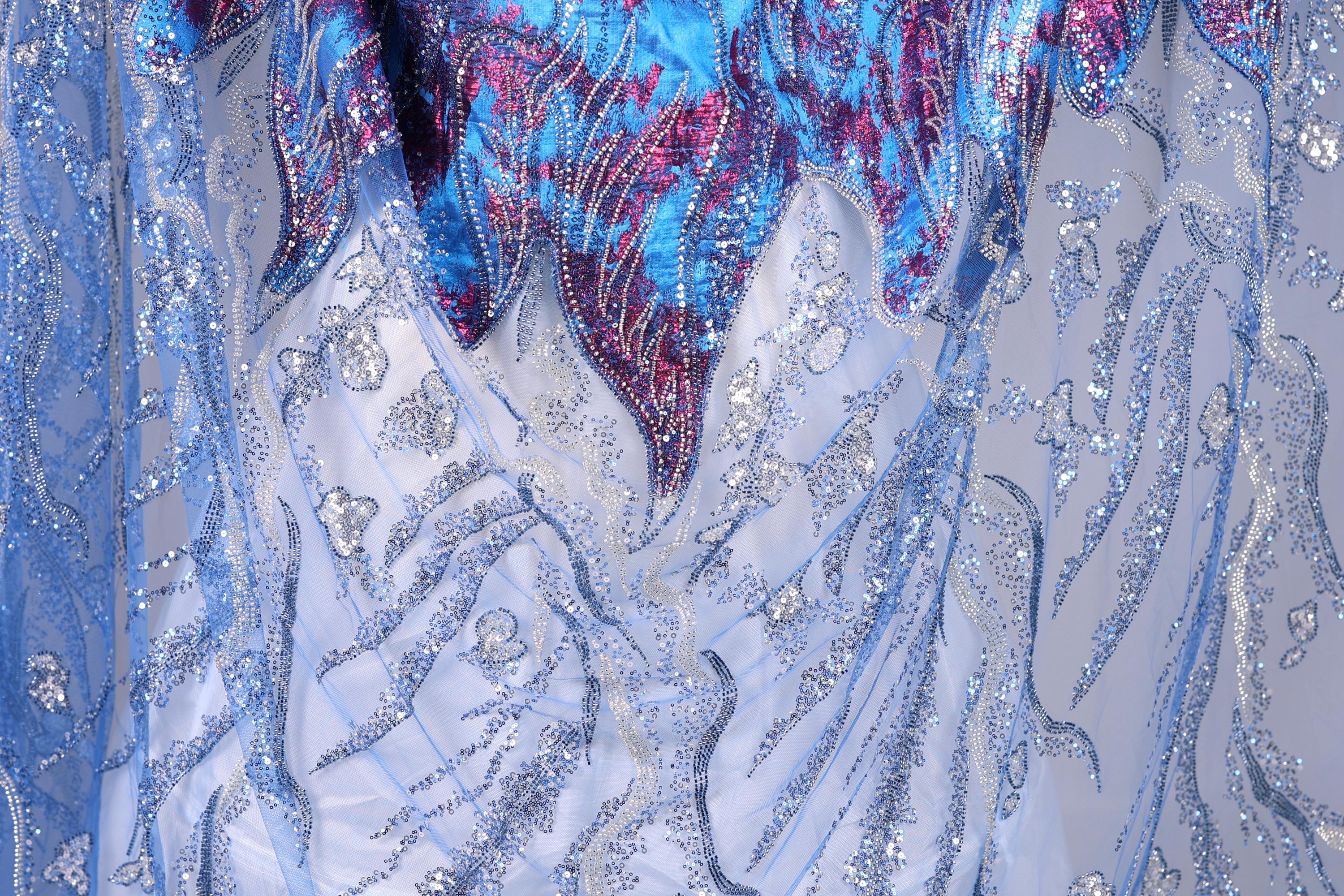 The image is a close-up view of a fabric with a blue and silver pattern, featuring a mix of metallic and floral elements. The fabric appears to be made of a shiny, sequined material, giving it a sparkling and luxurious appearance.