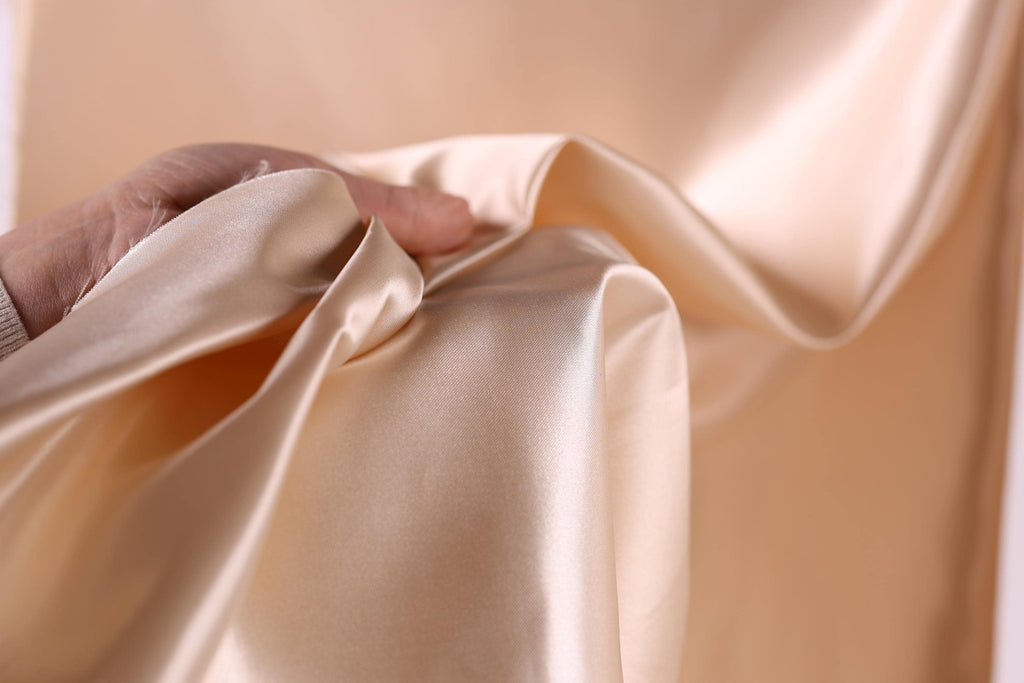 A hand is holding a piece of light-colored fabric, possibly silk or satin, in a close-up view.