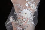 A close-up view of a delicate, white lace fabric adorned with intricate floral embroidery and sparkling rhinestones.
