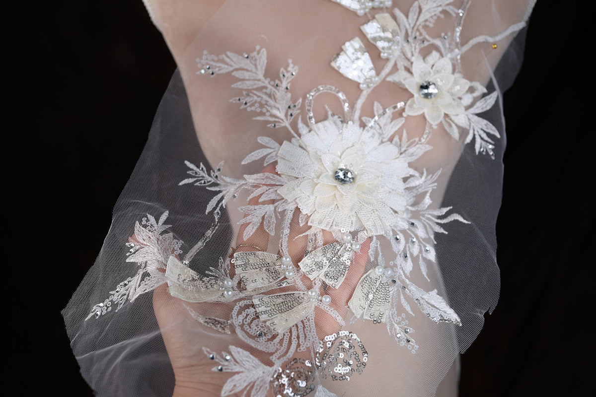 A close-up view of a delicate, white lace fabric adorned with intricate floral embroidery and sparkling rhinestones.