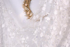 a close-up view of a delicate white lace dress with intricate floral embroidery. The dress is adorned with a wreath made of dried flowers, adding a touch of natural beauty to the overall design. The background is blurred, drawing attention to the details of the dress and the wreath.