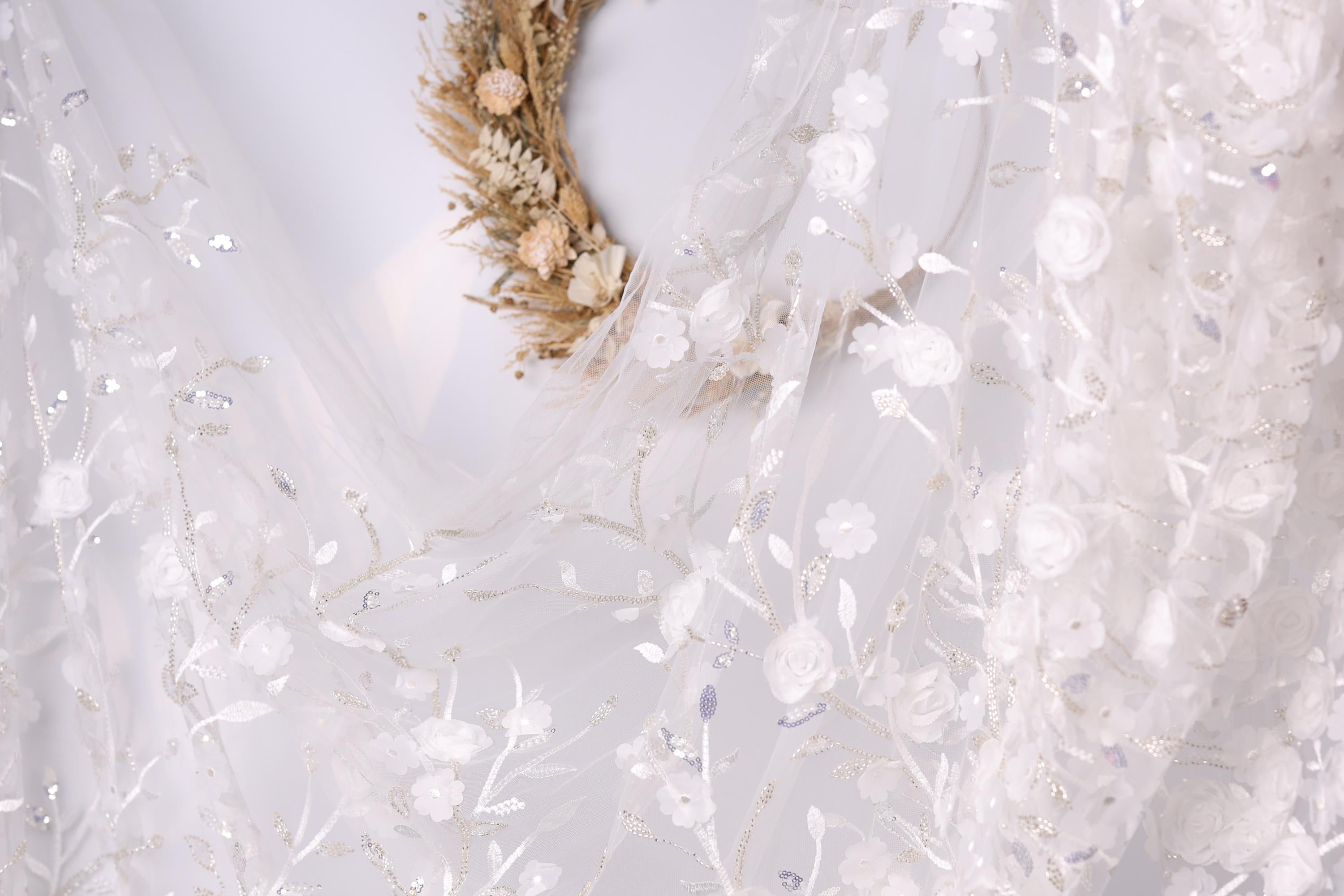 a close-up view of a delicate white lace dress with intricate floral embroidery. The dress is adorned with a wreath made of dried flowers, adding a touch of natural beauty to the overall design. The background is blurred, drawing attention to the details of the dress and the wreath.