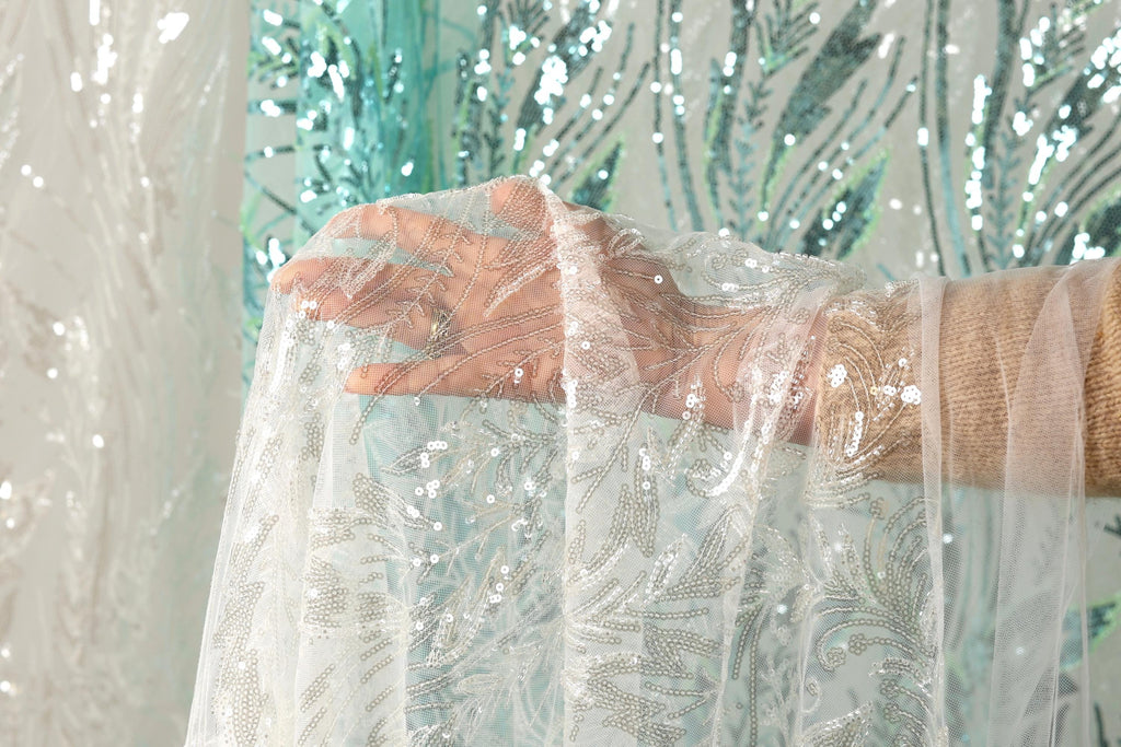 A hand is holding a piece of white lace with silver sequins, against a background of a green and white sequined curtain.