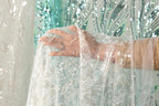 A hand is holding a piece of white lace with silver sequins, against a background of a green and white sequined curtain.