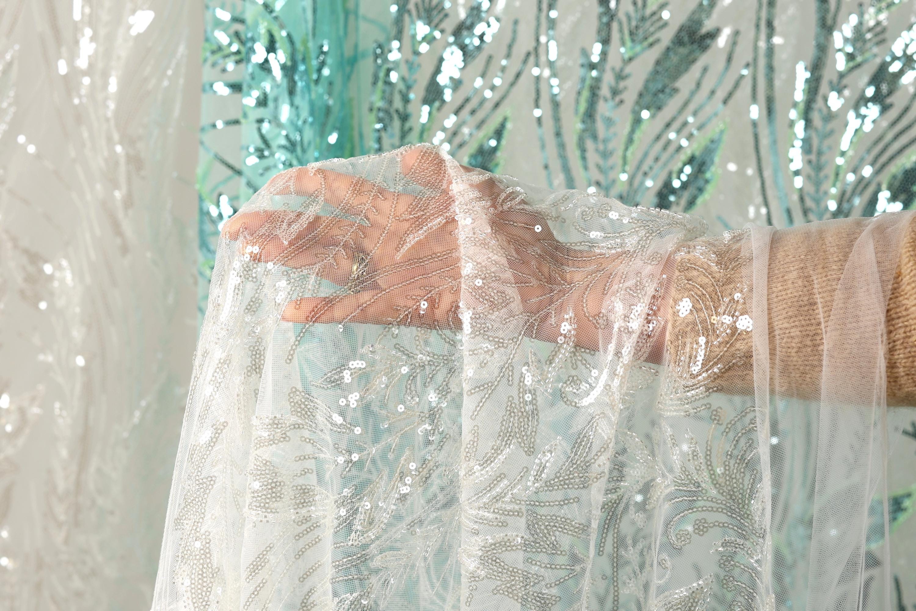 A hand is holding a piece of white lace with silver sequins, against a background of a green and white sequined curtain.