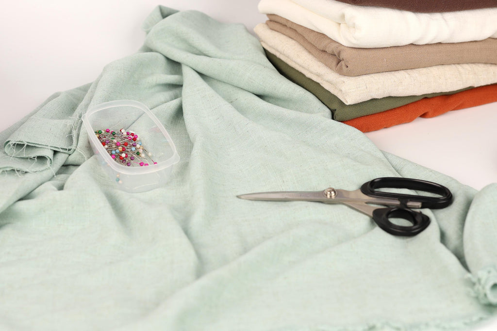 a pile of fabric, a pair of scissors, and a container of pins on a light green cloth.