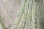 a close-up view of a delicate, intricately embroidered fabric with a floral pattern. The fabric appears to be made of a light green or mint-colored material, adorned with small, colorful beads or sequins that add a touch of sparkle and detail to the design. The image captures the fine details and textures of the fabric, showcasing the craftsmanship and artistry involved in its creation.