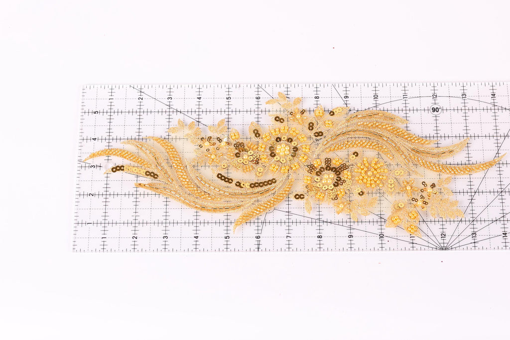 a decorative gold embroidered piece with intricate patterns and designs, including flowers and leaves, placed on a transparent ruler for scale.
