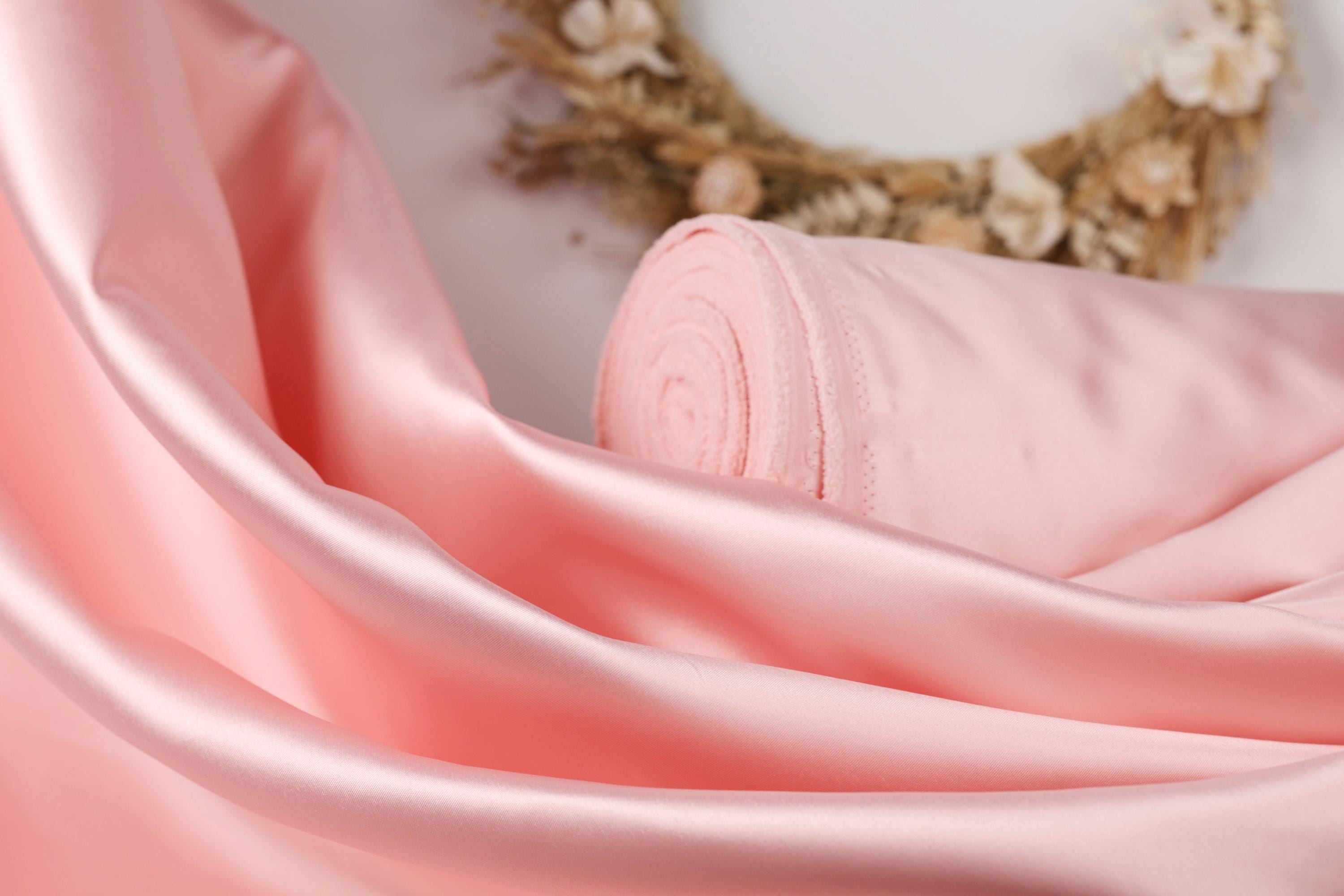 A close-up image of a soft, pink satin fabric, with a wreath of dried flowers visible in the background.