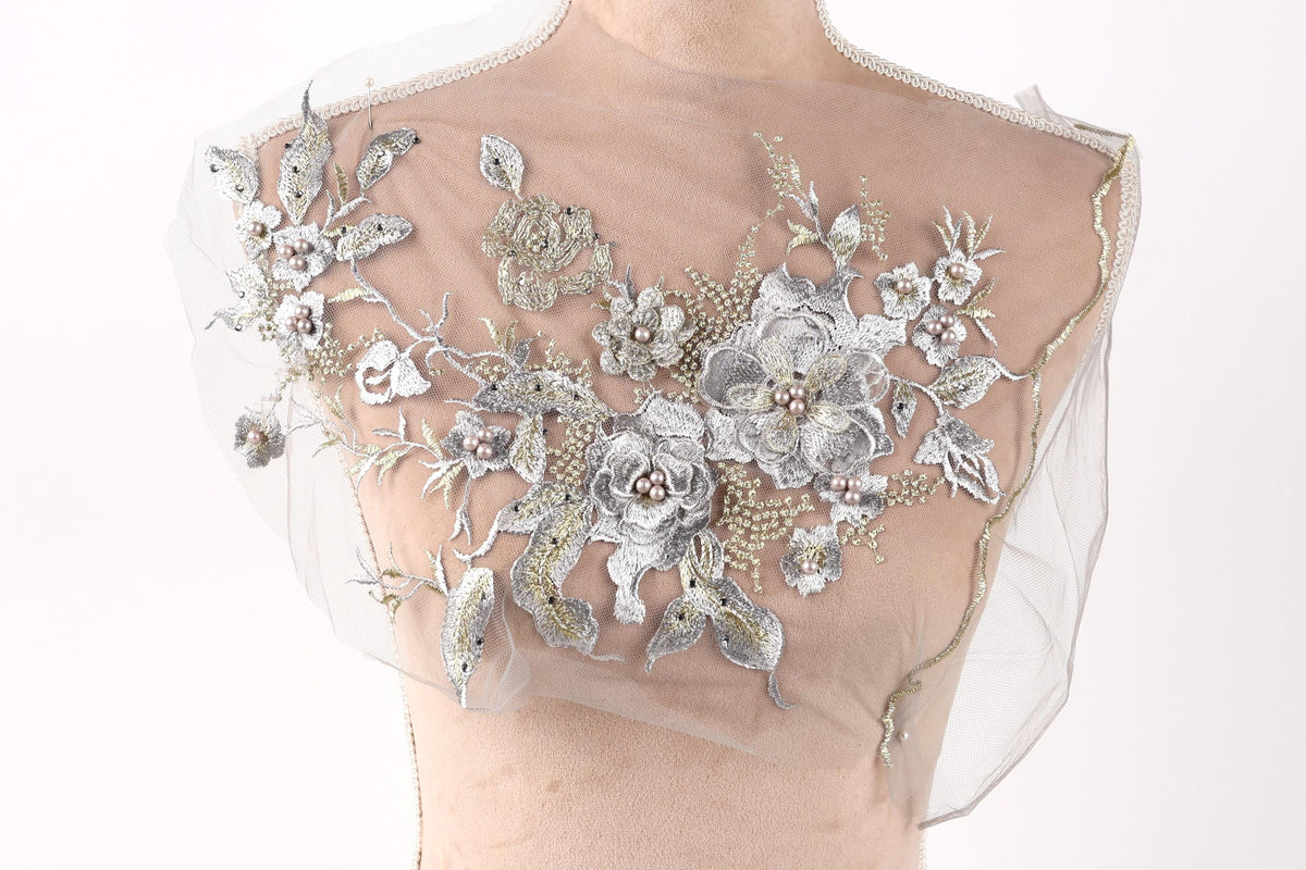 a mannequin with a delicate, intricate floral embroidery on its chest area. The embroidery is composed of silver and white flowers and leaves, creating a beautiful and elegant design. The mannequin is positioned against a plain white background, allowing the embroidery to stand out prominently.
