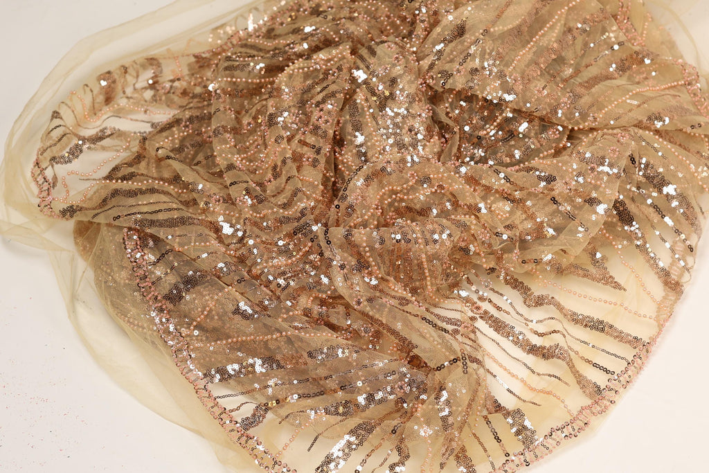 a close-up view of a piece of fabric with a gold and beige color scheme, adorned with numerous small sequins or beads that create a sparkling effect. The fabric appears to be made of sheer material, giving it a delicate and elegant appearance.