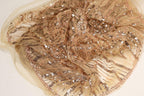 a close-up view of a piece of fabric with a gold and beige color scheme, adorned with numerous small sequins or beads that create a sparkling effect. The fabric appears to be made of sheer material, giving it a delicate and elegant appearance.