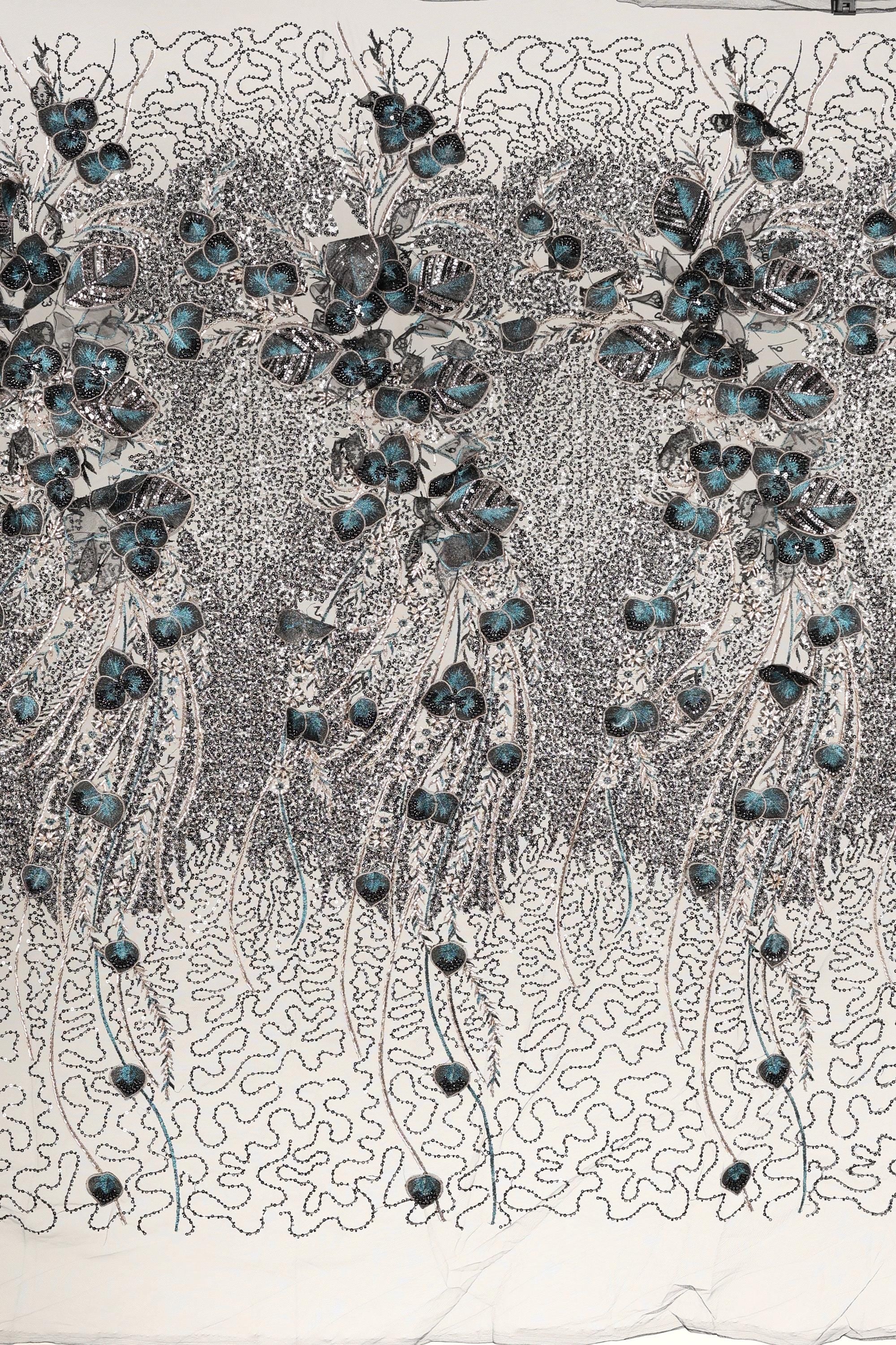 The image is a close-up of a fabric with a floral pattern, featuring a repeating design of blue flowers and leaves. The fabric appears to be made of a material with a textured surface, and the pattern is intricate and detailed.