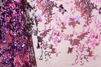 a close-up view of a fabric with a floral pattern in shades of pink and purple. The fabric appears to be a sheer material, with a mix of sequins and lace-like elements. The pattern is intricate and detailed, showcasing the craftsmanship of the fabric.