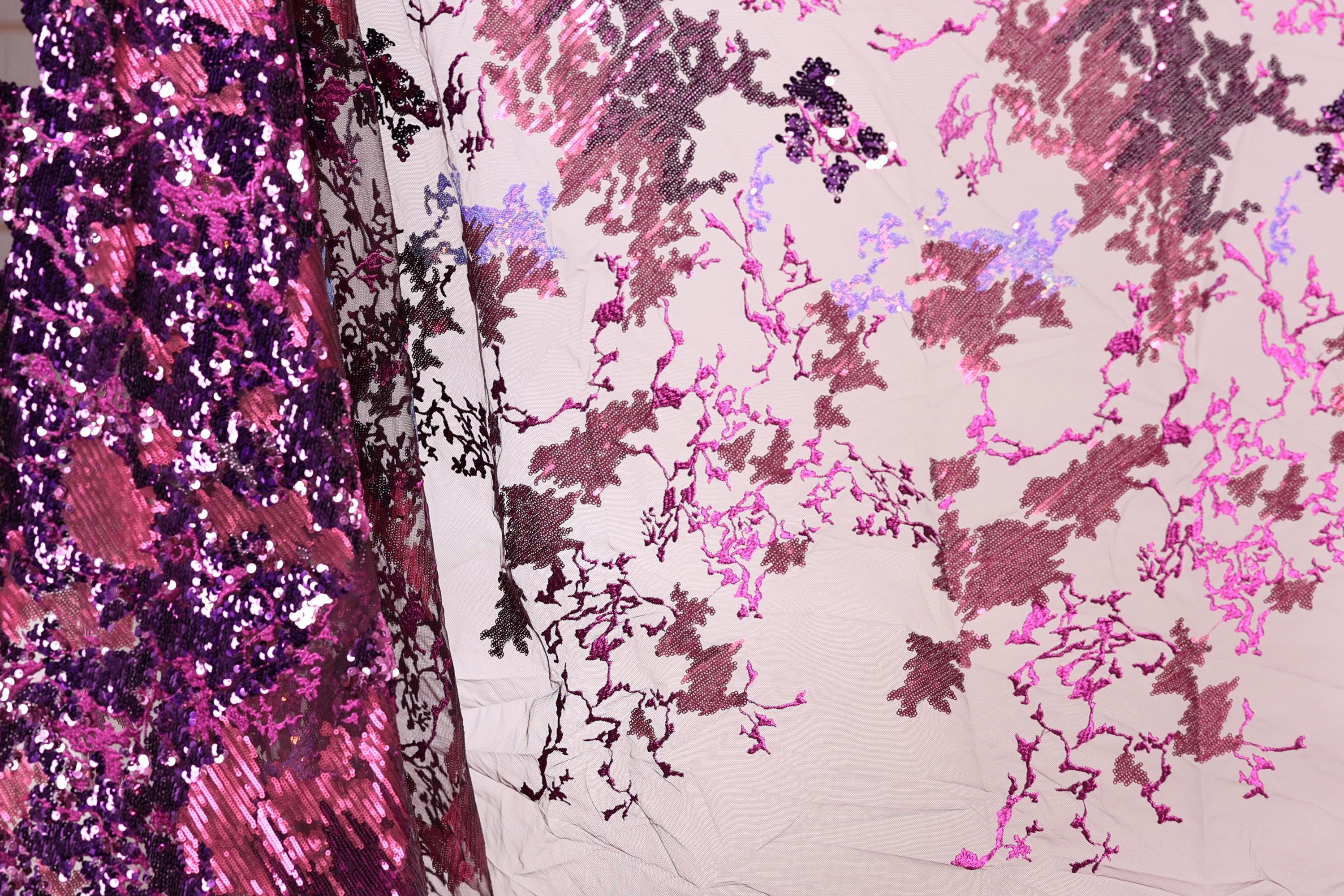 a close-up view of a fabric with a floral pattern in shades of pink and purple. The fabric appears to be a sheer material, with a mix of sequins and lace-like elements. The pattern is intricate and detailed, showcasing the craftsmanship of the fabric.