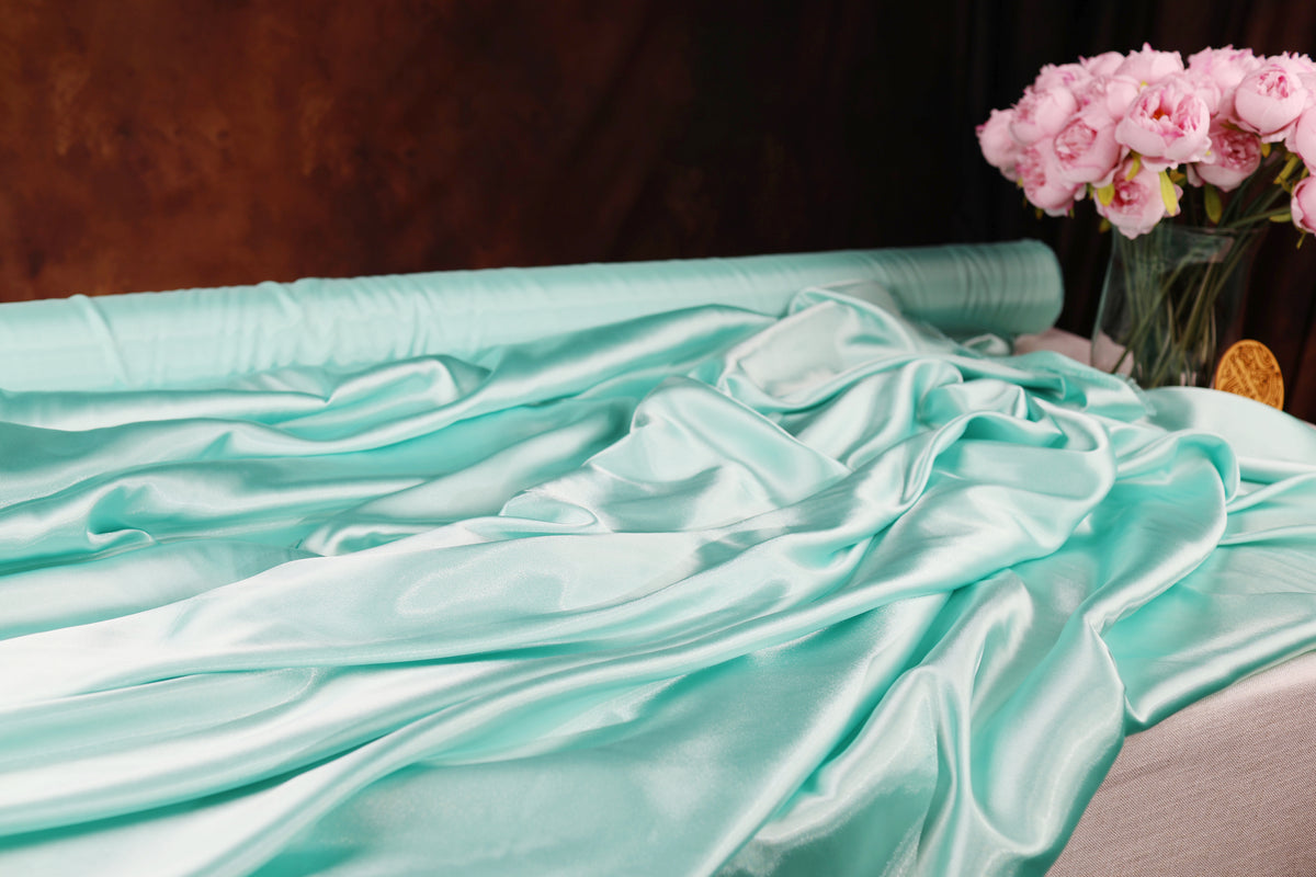 a close-up view of a turquoise satin fabric draped over a surface, with a vase of pink flowers placed next to it.