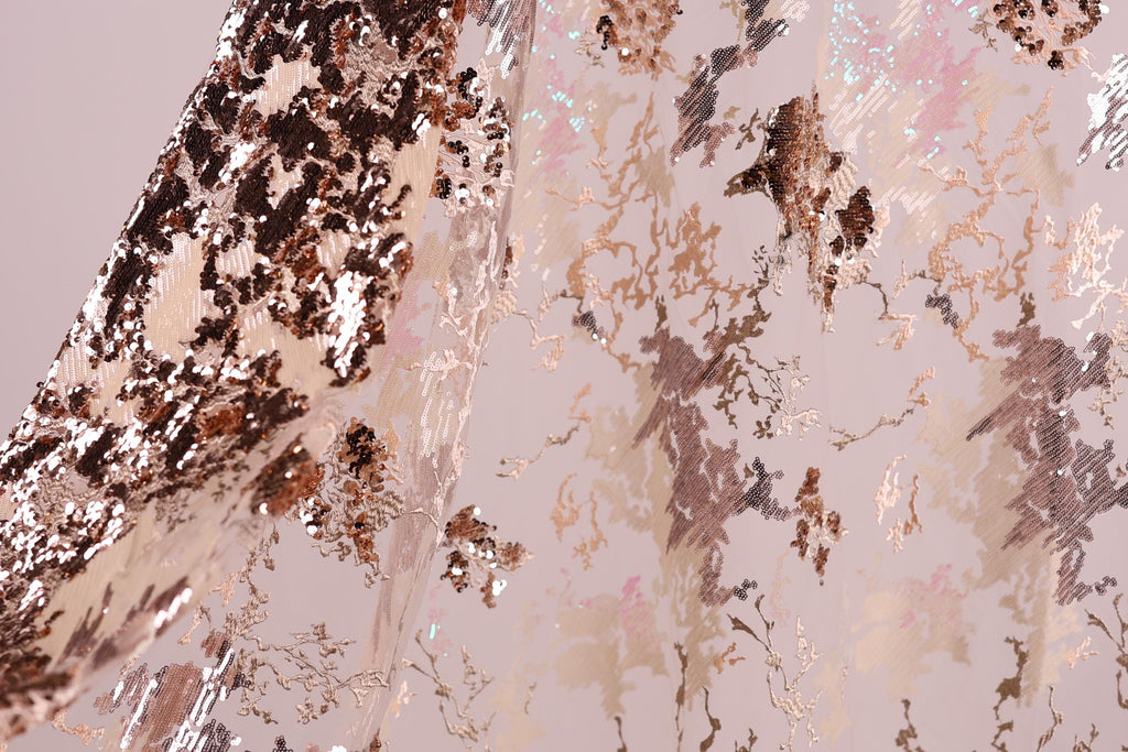 a close-up view of a fabric with a metallic, sequined texture. The fabric appears to be a light pink color, with a pattern of small, irregularly shaped sequins or glittering particles covering the surface. The fabric is folded or crumpled, creating a sense of depth and texture in the image.