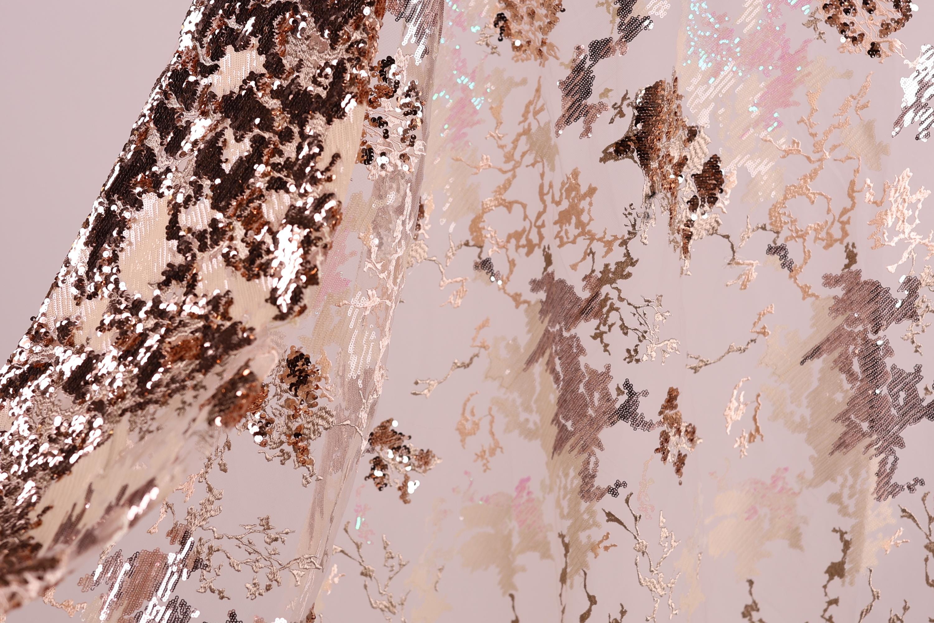 a close-up view of a fabric with a metallic, sequined texture. The fabric appears to be a light pink color, with a pattern of small, irregularly shaped sequins or glittering particles covering the surface. The fabric is folded or crumpled, creating a sense of depth and texture in the image.