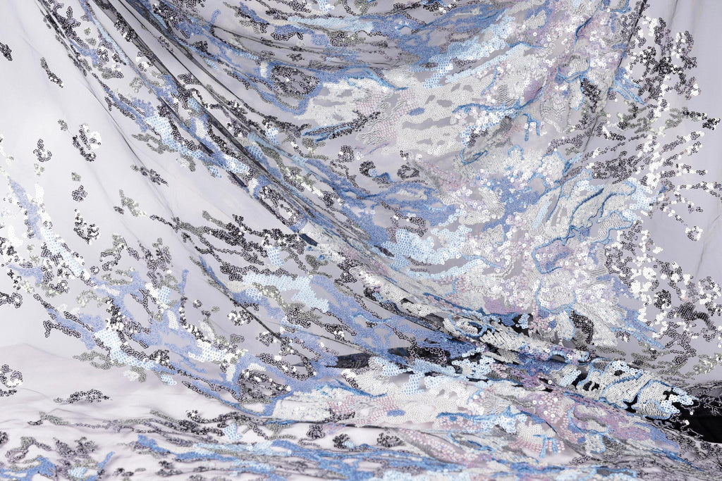 The image is a close-up of a fabric with a complex, swirling pattern of sequins in shades of blue, gray, and white. The fabric appears to be made of sheer material, giving it a delicate and ethereal appearance. The intricate design and the interplay of the different colors create a visually striking and captivating image.