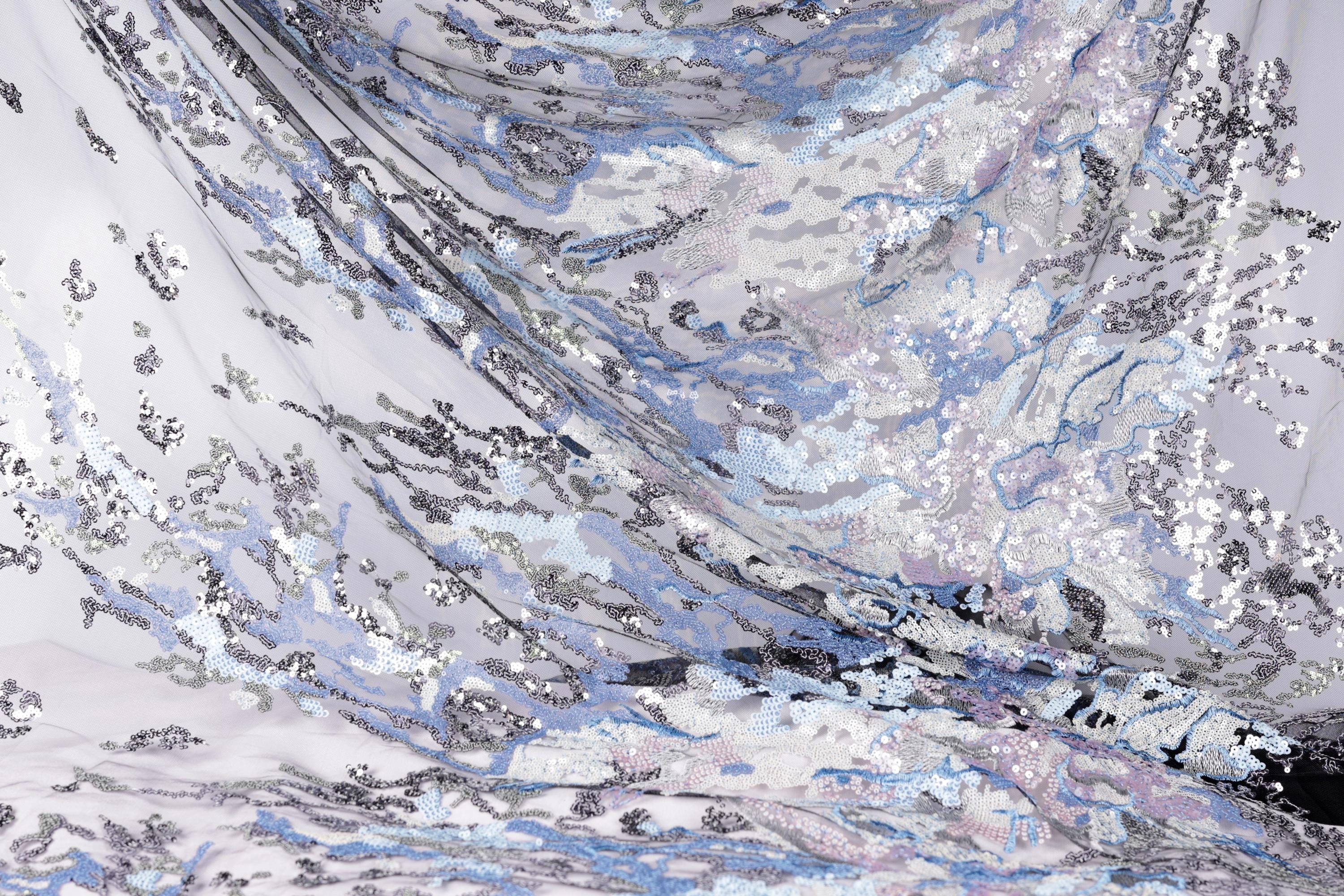 The image is a close-up of a fabric with a complex, swirling pattern of sequins in shades of blue, gray, and white. The fabric appears to be made of sheer material, giving it a delicate and ethereal appearance. The intricate design and the interplay of the different colors create a visually striking and captivating image.