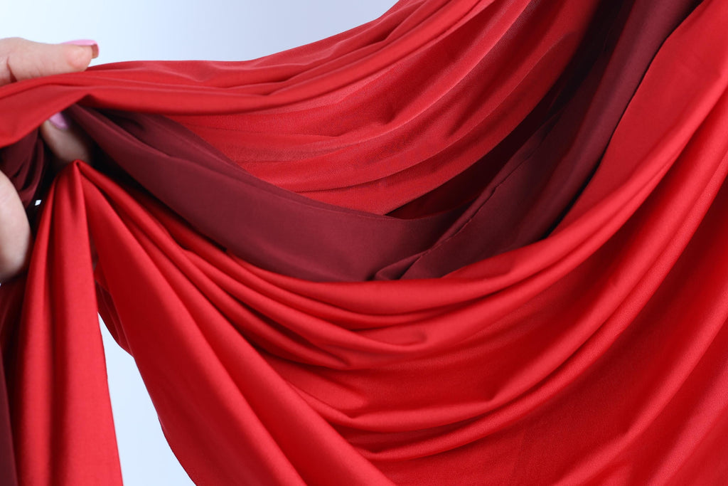A hand is holding a red fabric, which appears to be a sheer or sheer-like material. The fabric is folded and draped in a way that creates a sense of movement and texture.