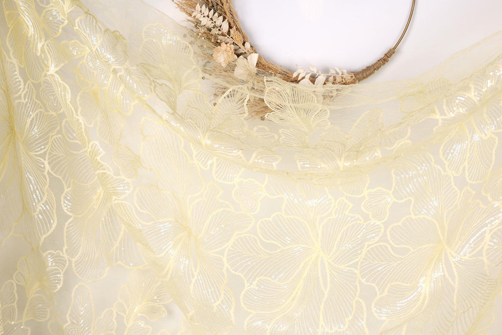 a close-up view of a delicate, yellow fabric with a floral pattern. The fabric appears to be made of sheer material, with a delicate and intricate design. The background is plain white, which allows the fabric and its details to stand out prominently.