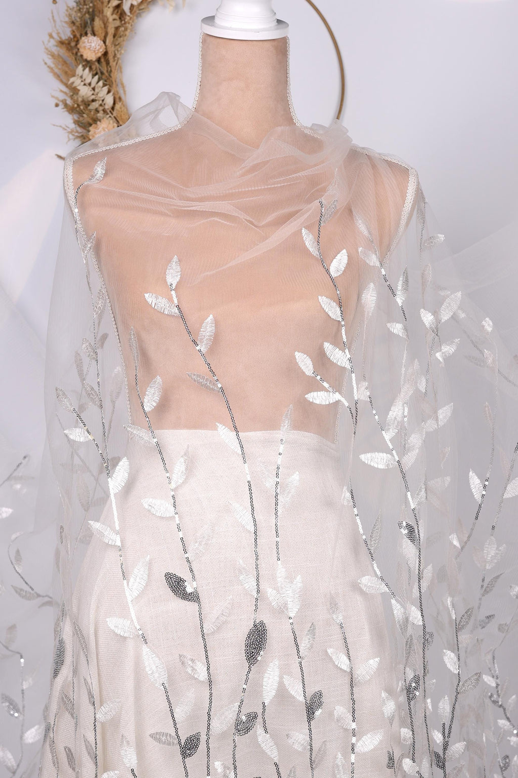 A mannequin wearing a white dress with a sheer, leaf-like fabric draped over it.