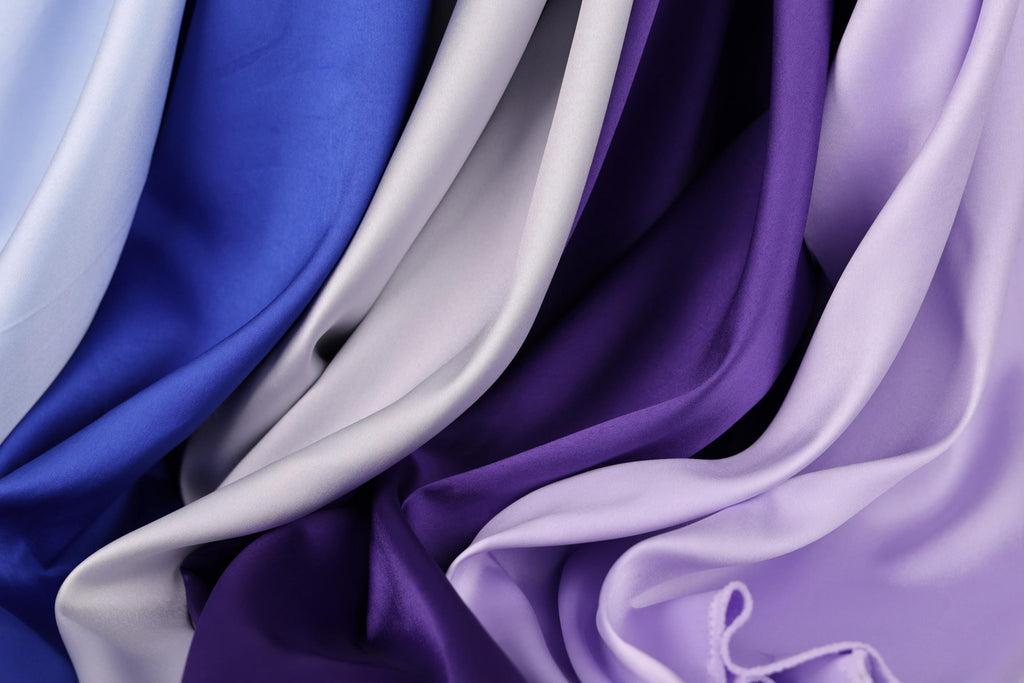a close-up view of various colored satin fabrics, including blue, gray, and purple, arranged in a crumpled and flowing manner.