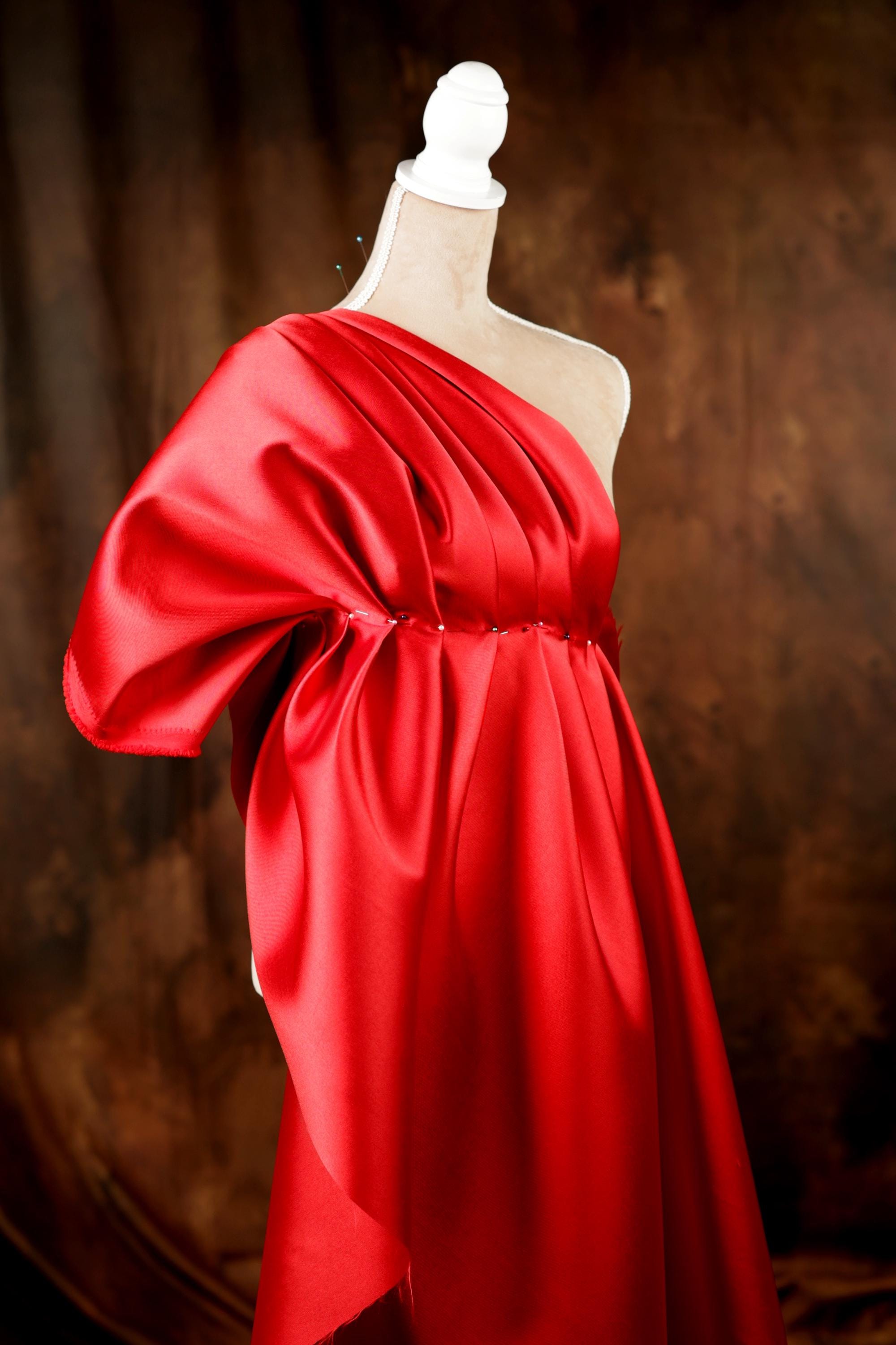 A red one-shoulder dress with a draped, flowing silhouette is displayed on a mannequin against a dark background.