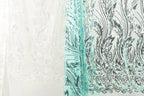a close-up view of two fabric pieces with intricate patterns. The fabric on the left is white, while the fabric on the right is teal. The patterns on both fabrics are similar, with a floral or leaf-like design. The image does not contain any text or additional objects, and the focus is solely on the two fabric pieces.
