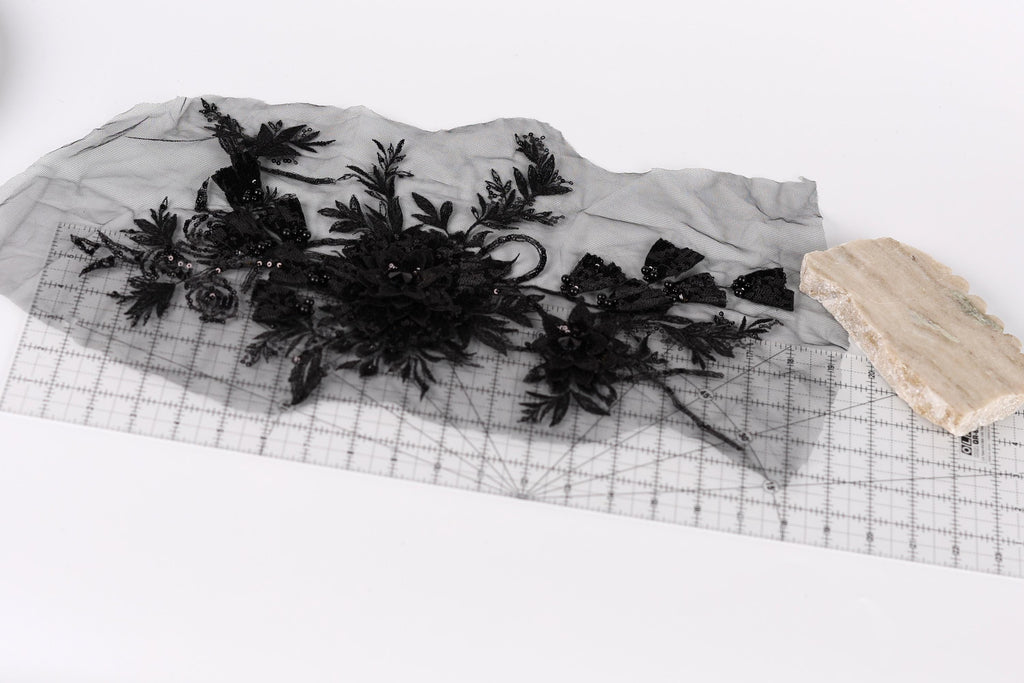 a piece of black fabric with intricate floral designs, placed on a transparent surface. Next to the fabric, there is a small piece of light-colored material, possibly a piece of paper or a small object.