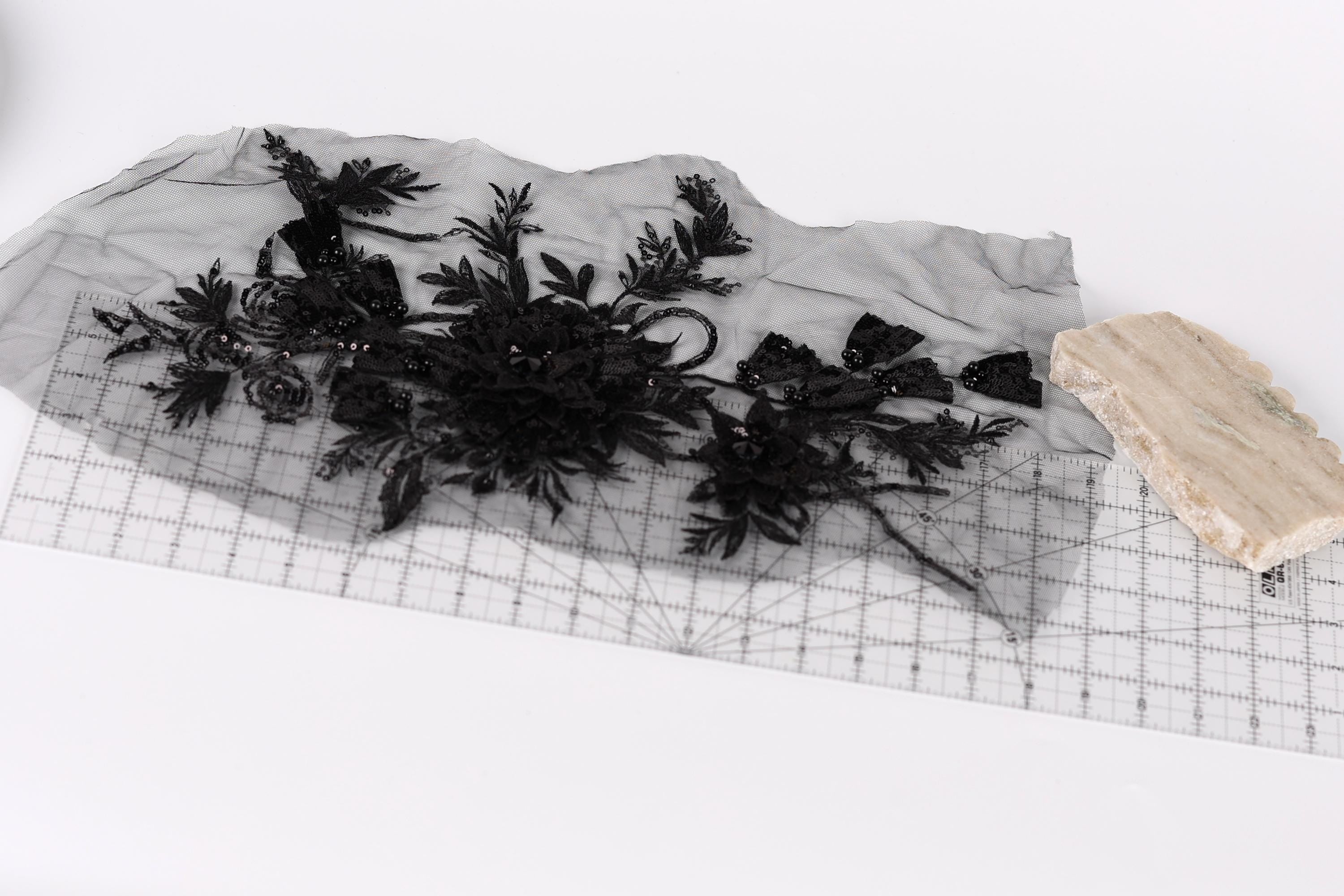 a piece of black fabric with intricate floral designs, placed on a transparent surface. Next to the fabric, there is a small piece of light-colored material, possibly a piece of paper or a small object.