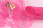 A glass vase filled with flowers sits on a pink background, accompanied by a roll of pink fabric.