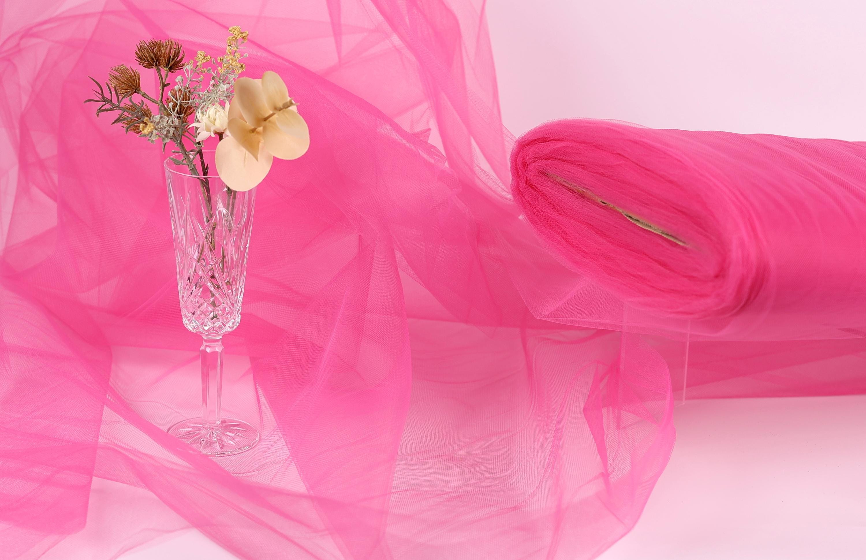 A glass vase filled with flowers sits on a pink background, accompanied by a roll of pink fabric.