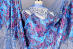 a close-up view of a blue and purple patterned fabric, possibly a dress or garment. The fabric has a floral or paisley design, with a mix of blue, purple, and pink colors. The fabric appears to be made of a shiny, sequined material, adding a touch of sparkle and elegance to the design.